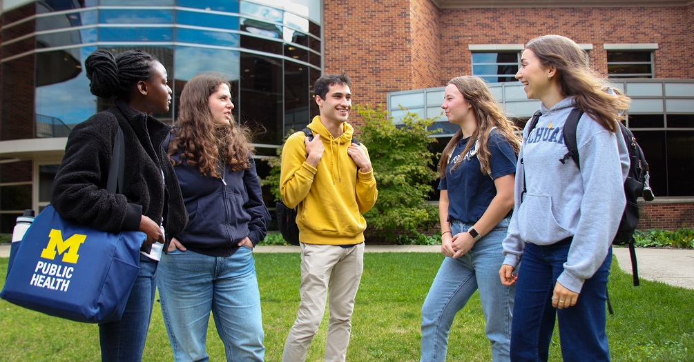.Five diverse University of Michigan public health undergraduate students talking and laughing together on campus lawn in front of modern brick academic building, with one student carrying a blue Michigan Public Health tote bag