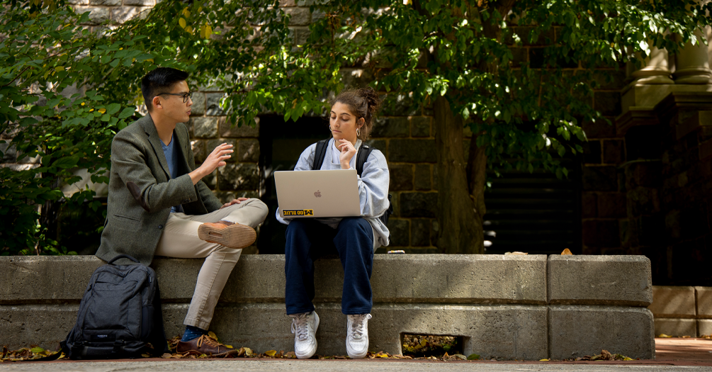 Two students sit on a concrete bench in the shade discussing their classwork at the U-M Ann Arbor campus