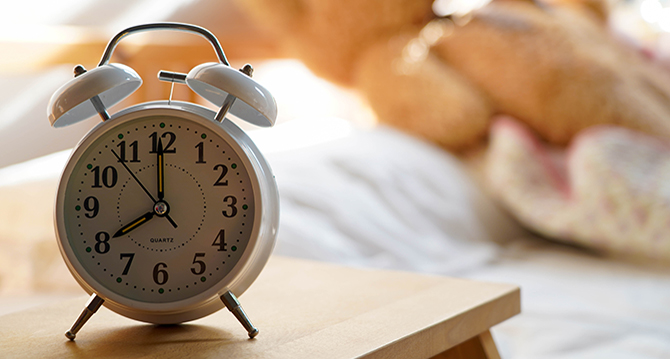A clock in the foreground and a teddy bear on a bed in the background.