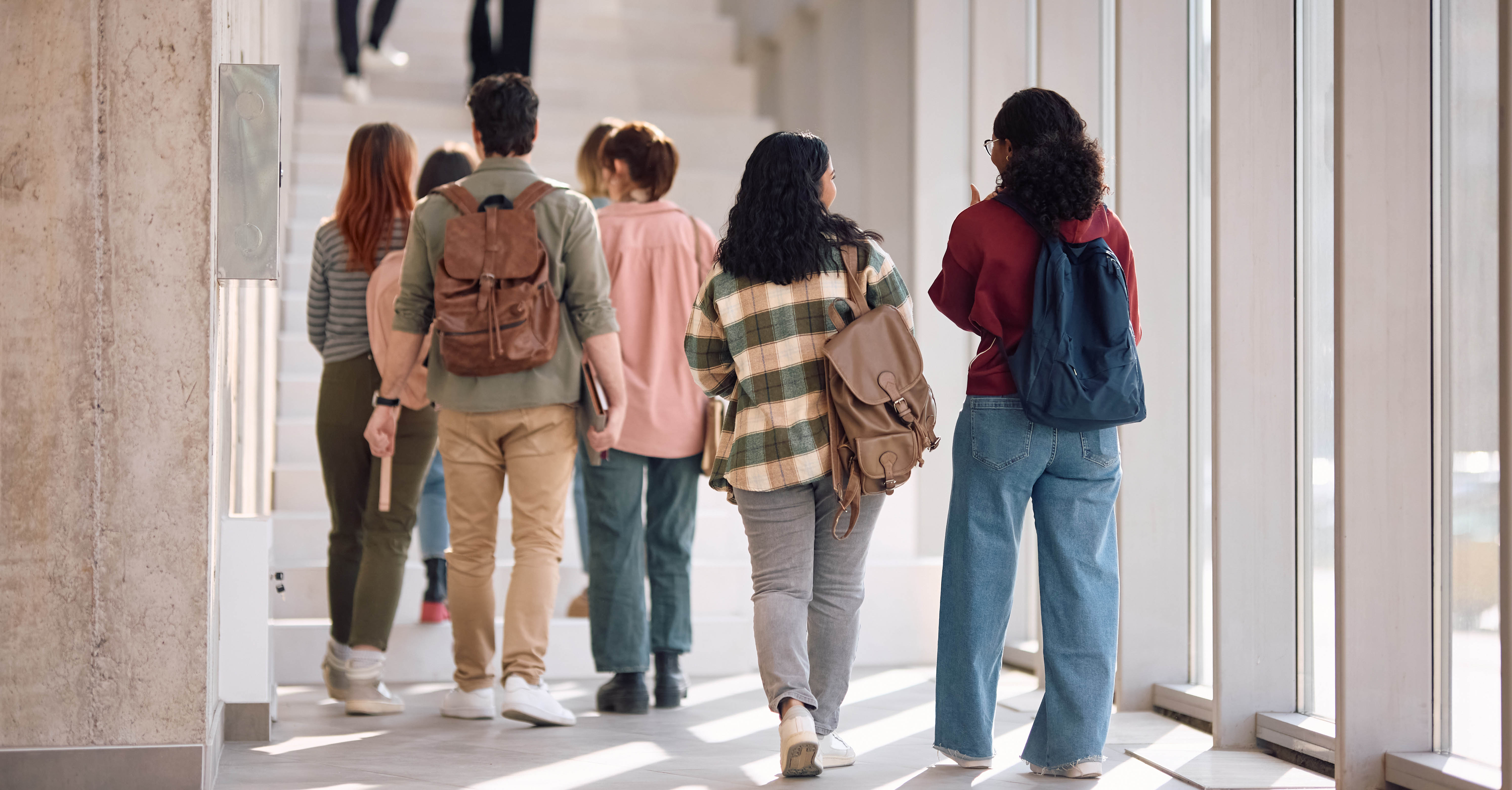 A group of people walking through a brightly lit corridor, viewed from behind. They are casually dressed and carry backpacks.