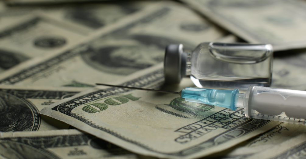 A vaccine needle and vial on a stack of hundred dollar bills.