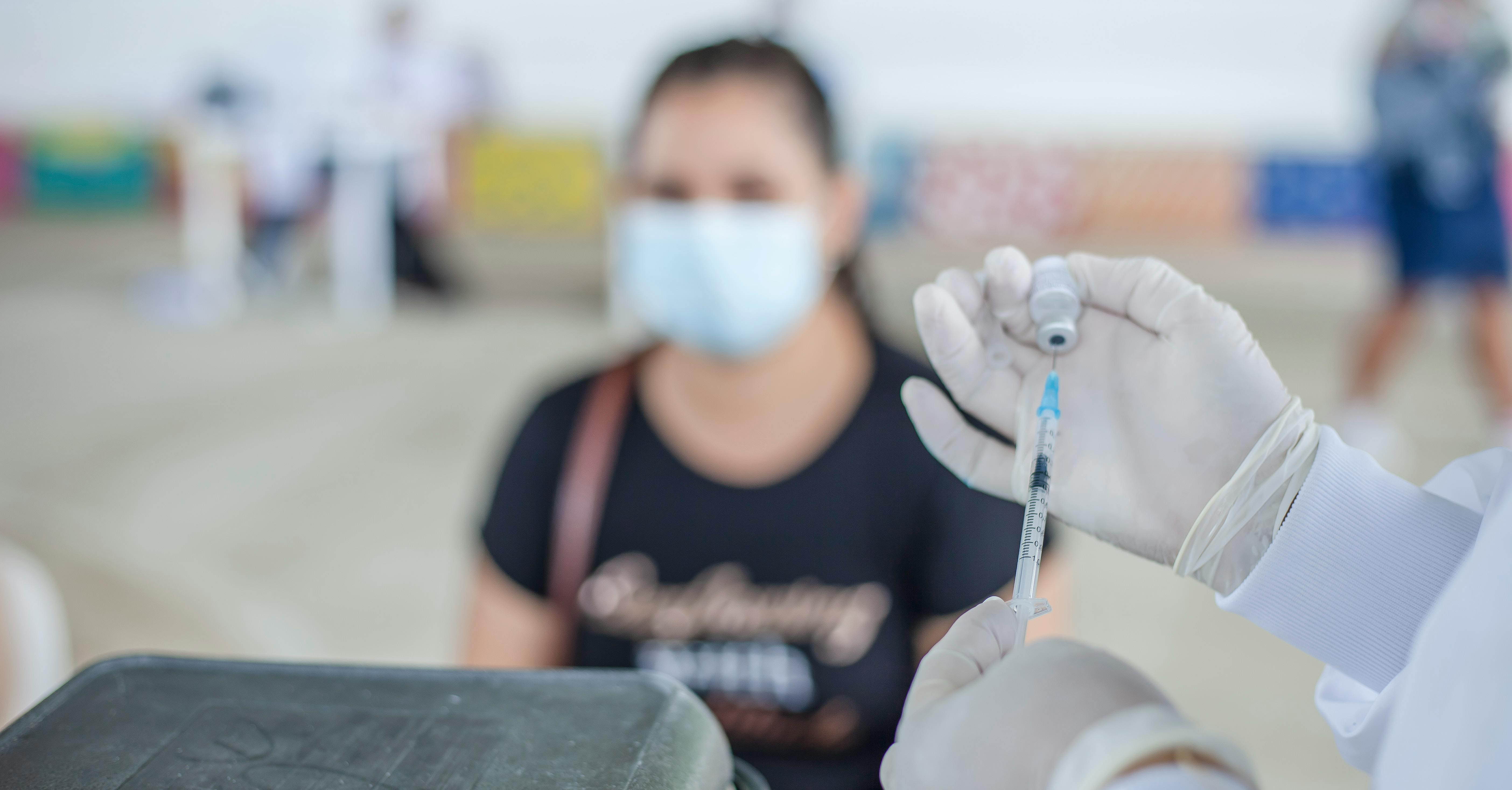 A person holding a syringe to administer a vaccine. In the background, A patient sits in a chair. 