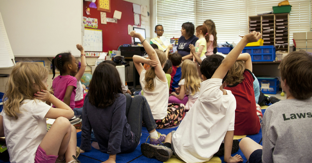A photo of a classroom, with a large group of elementary school students sitting on a carpet. Some are raising their hands. In the background, out of focus, three children stand with the teacher.