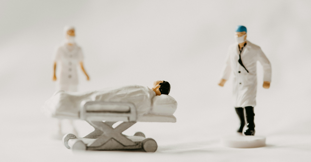 A blurry photo of three figurines: a patient in a hospital bed, a nurse, and a doctor. In front of a white background